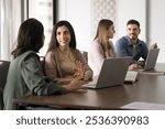 Positive beautiful young Latin professional woman talking to colleague at workplace. Hispanic business coworkers meeting at work table, discussing project tasks, teamwork, cooperation strategy