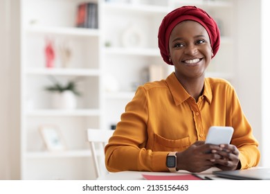 Positive beautiful young muslim black woman in red headscarf using brand new mobile phone, looking at copy space and smiling, using newest mobile application for business, home interior