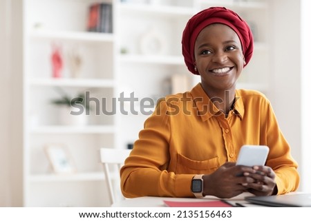 Positive beautiful young muslim black woman in red headscarf using brand new mobile phone, looking at copy space and smiling, using newest mobile application for business, home interior Foto stock © 