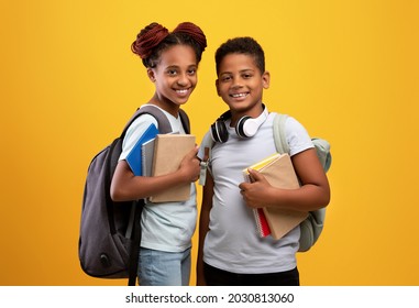Positive black boy and girl schoolers with backpacks holding books and notepads posing on yellow studio background, cheerful afro-american brother and sister ready to go to school