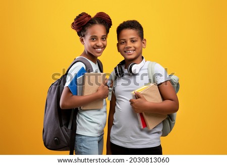 Positive black boy and girl schoolers with backpacks holding books and notepads posing on yellow studio background, cheerful afro-american brother and sister ready to go to school