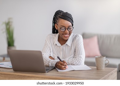 Positive black business woman with stylish glasses manager working from home during COVID-19 pandemic, using modern laptop and taking notes, sitting at workdesk in living room, copy space