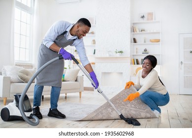 Positive black couple cleaning up together, vacuuming under carpet at their house, copy space