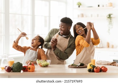 Positive Black Family Of Three Cooking And Having Fun Singing Favorite Song Holding Spoons Preparing Dinner Together Standing In Modern Kitchen Indoor. Weekend Leisure, Healthy Food Recipes Concept