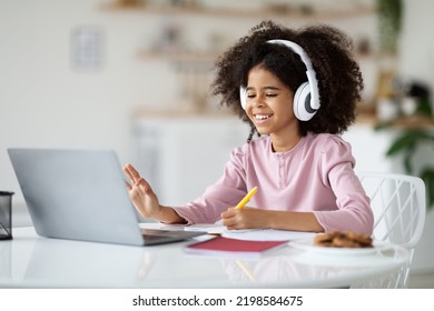 Positive Black Kid School Girl With Bushy Hair Having Online Party With Friends While Staying Home, Sitting At Desk, Drawing, Using Wireless Headset, Waving And Smiling At Computer Screen, Copy Space