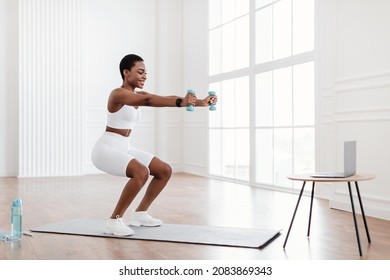 Positive Black Lady Exercising And Squatting With Two Dumbbells In Front Of Pc On Tea Table At Home, Smiling Fitness Blogger Recording Video For Her Followers Having Personal Virtual Training