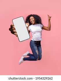 Positive black lady showing smartphone while jumping in air over pink studio background, mockup for app or website design on white screen. Creative collage with advertising template