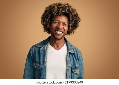 Positive black male in denim jacket and white t shirt with curly hair smiling and looking at camera against brown background - Powered by Shutterstock