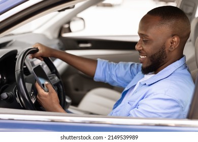 Positive black male driver holding mobile phone while driving nice auto, using new mobile application for navigation going on vacation or making summer trip, stuck in traffic jam, profile side view