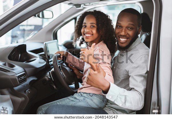 Positive\
black man and his cute daughter sitting on driver seat of new car,\
showing thumbs up at auto dealership. Friendly African American\
family test driving vehicle together at\
showroom