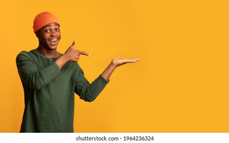 Positive Black Man Pointing Finger At Open Palm, Posing Isolated On Yellow Background, Cheerful African Guy In Stylish Hat Advertising Something, Showing Invisible Object, Panorama With Copy Space