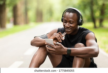 Positive black sportsman eating protein bar and using phone while resting, exercising at park, empty space - Powered by Shutterstock