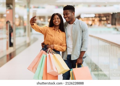 Positive Black Spouses Shopping In Modern City Mall And Taking Selfie On Smartphone, African American Couple Having Fun, Enjoying Doing Purchases Together, Carrying Shopper Bags, Blurred Background