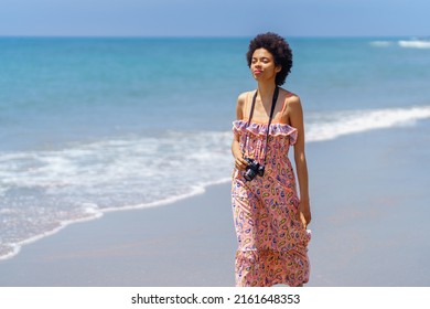 Positive Black Woman Walking On The Sand Of A Tropical Beach In A Summer Dress And Slr Camera. Girl Enjoying Her Holiday In A Coastal Area.