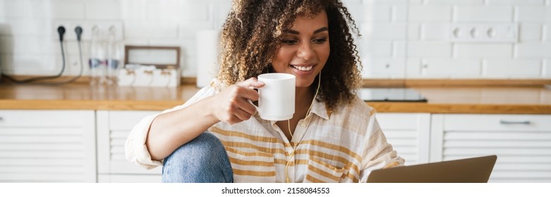 A positive black woman in a wired headphones with a cup in her hands looking at the laptop screen while sitting in the white kitchen - Shutterstock ID 2158084553