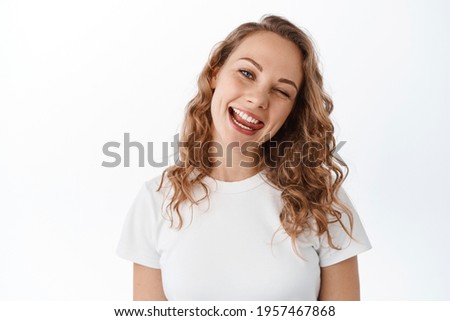 Positive blond girl winking, showing tongue and smiling happy, staying upbeat and optimistic, looking with joy at camera, standing in t-shirt against white background