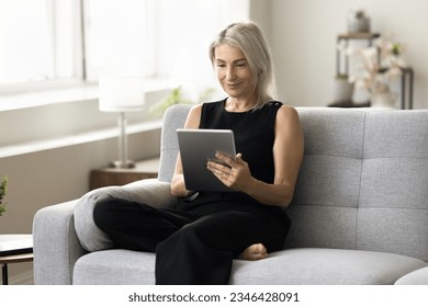 Positive blonde mature woman using online application on tablet computer at home, relaxing on couch, smiling, reading book on Internet, enjoying wireless Internet technology on digital device - Powered by Shutterstock