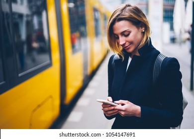 Positive blonde woman holding cellular updating information about city transport on web page, smiling hipster girl satisfied with online ticket service paying for electric transport via smartphone