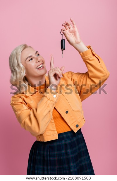 Positive blonde woman pointing at car key isolated\
on pink