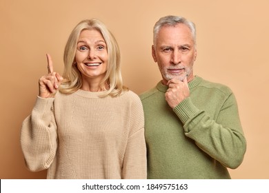 Positive blonde woman points above with glad expression wears casual jumper. Serious bearded man holds chin tries to think over something. Married couple enjoy their retirement isolated on brown wall