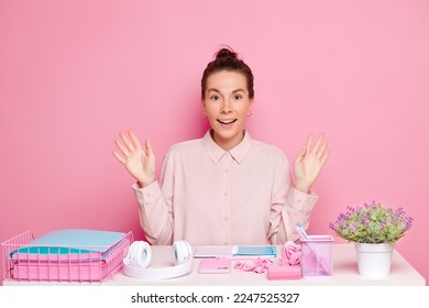 Positive brunette adult woman in formal shirt shrugs shoulders   spread palms aside  smiles widely  feels excited during creative task in office  poses over pink wall