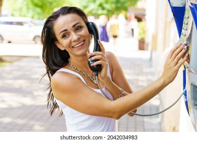 Positive brunette woman holding telephone receiver calls through stationery phone in street, tourist talking on payphone working with coins, happy with international communication on sunny summer day