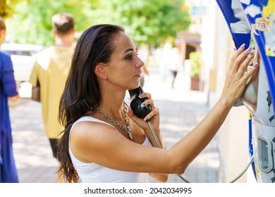 Positive brunette woman holding telephone receiver calls through stationery phone in street, tourist talking on payphone working with coins, happy with international communication on sunny summer day