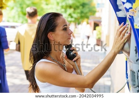 Positive brunette woman holding telephone receiver calls through stationery phone in street, tourist talking on payphone working with coins, happy with international communication on sunny summer day