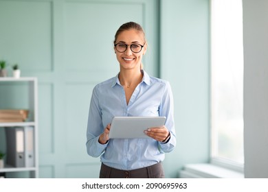 Positive Business Lady In Formal Wear Using Tablet In Office, Looking And Smiling At Camera. Successful Female Entrepreneur Checking Email, Standing Near Workplace