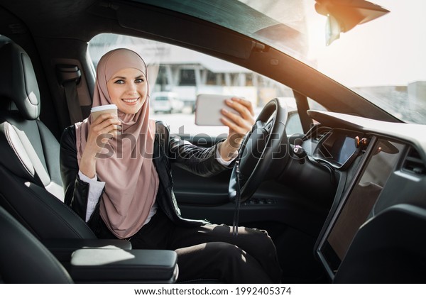 Positive business woman in\
hijab having video call on smartphone while sitting in luxury\
electric car. Attractive lady drinking fresh coffee and talking on\
mobile.