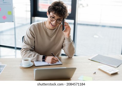 Positive businessman talking on mobile phone and writing on notebook near laptop, coffee and calculator in office - Powered by Shutterstock