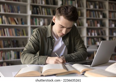 Positive busy hardworking student guy writing draft, notes, article, essay, webinar summary, studying in college library, working on research at desk with open books, laptop computer