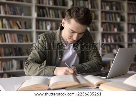 Positive busy hardworking student guy writing draft, notes, article, essay, webinar summary, studying in college library, working on research at desk with open books, laptop computer