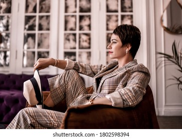 Positive And Calm Short Haired Brunette Woman In Stylish Business Plaid Pantsuit With Rolled Up Sleeves Sits In Armchair  Holding Hat In Hand In Room. Business Elegance Concept