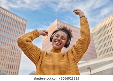 Positive carefree curly haired woman dances with joy enjoys favorite audio track via headphones dressed in casual knitted jumper poses against skyscrapers and blue sky spends free time at city