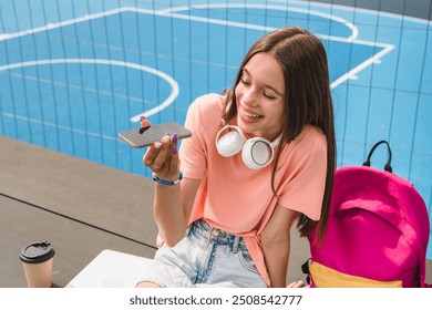 Positive carefree high school girl teenager pupil college student in casual clothes with wireless headphone holding using mobile phone recording voice meassage sitting in sport court yard - Powered by Shutterstock