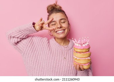 Positive carefree woman holds tasty sweet ccandles with burning candles smiles broadly makes peace sign over eye enjoys festive event and celebration wears casual knitted sweater isolated on pink wall