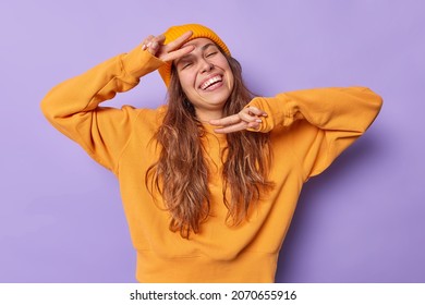 Positive carefree woman makes peace gesture or v sign smiles happily enjoys moment dressed in casual clothes poses against purple background. People positive emotions and body language concept - Shutterstock ID 2070655916