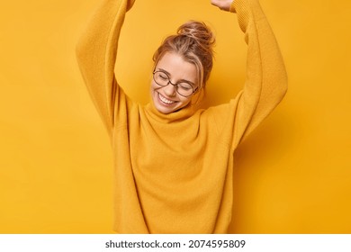 Positive carefree woman raises arms up dances carefree from joy wears round transparent glasses and jumper smiles gladfully keeps eyes closed isolated over vivid yellow background rejoices success - Shutterstock ID 2074595809