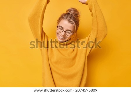 Positive carefree woman raises arms up dances carefree from joy wears round transparent glasses and jumper smiles gladfully keeps eyes closed isolated over vivid yellow background rejoices success