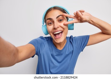 Positive carefree woman takes selfie makes peace gesture dressed in casual blue t shirt listens music via stereo headphones isolated over white background. Glad female meloman enjoys favorite playlist