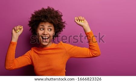 Positive carefree young woman with Afro hairstyle feels relaxed and happy, has fun at party, gazes aside, has beaming smile, isolated on purple background. People, hapiness, lifestyle concept
