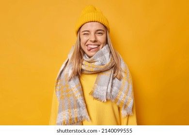 Positive carefree young woman winks eye and sticks out tongue wears hat casual jumper scarf around neck foolishes around isolated over yellow background goes crazy. Human facial expressions.