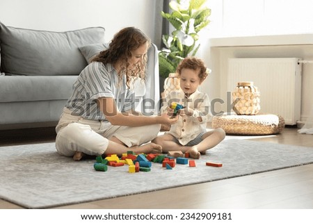 Positive caring young mom and cute toddler kid playing toy building blocks on carpeted floor, sitting at heap of colorful cubes in home living room. Mother teaching child, helping to improve skills