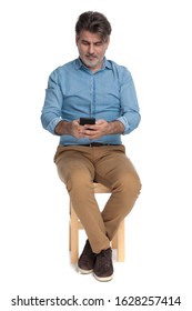 Positive Casual Man Writing On His Phone And Smiling While Sitting On A Chair On White Studio Background