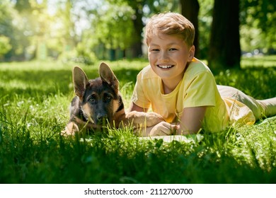 Positive Caucasian Child In Summer Outfit Relaxing With Cute Puppy At Green City Park. Happy Male Owner Enjoying Time Spending With Lovely Pet.