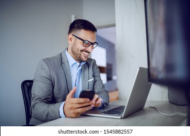 Positive Caucasian Elegant Businessman In Suit And With Eyeglasses Holding Smart Phone While Looking At Laptop. Office Interior.