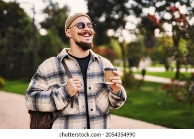 Positive Caucasian Hipster Bearded Guy With Mustache In Hat And Black Sunglasses Holds Coffee To Go While Walks In Urban Setting With Backpack 