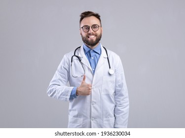 Positive Caucasian male doctor in lab coat showing thumb up gesture over grey studio background - Powered by Shutterstock