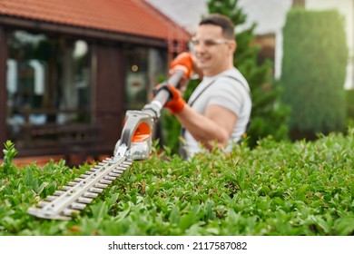 Positive caucasian man in safety glasses and gloves cutting bushes on backyard. Competent gardener using modern electric trimmer for work outdoors.